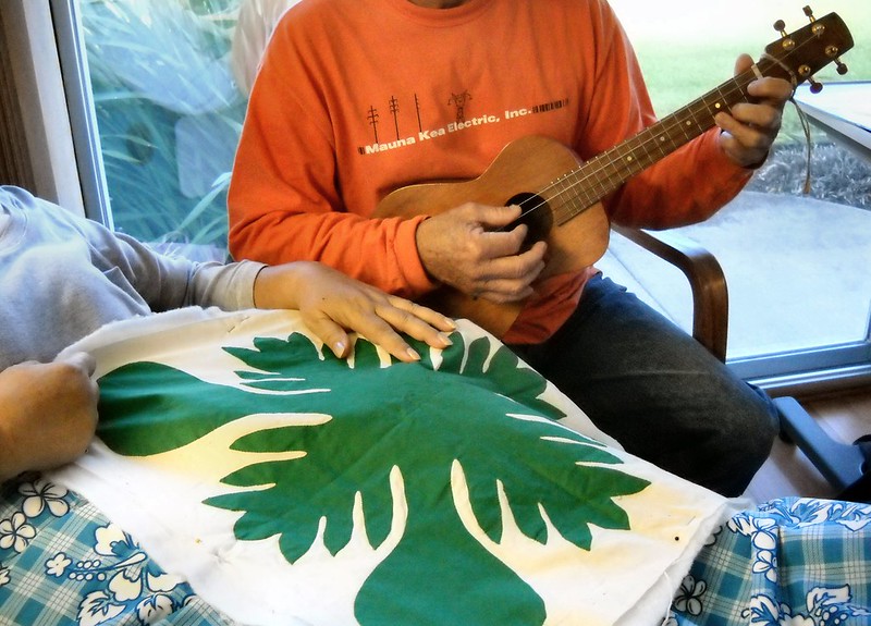 person working on Hawaiian quilt while another strums an ukulele